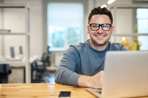 A man in a grey sweater wearing glasses sits at a desk behind a laptop.